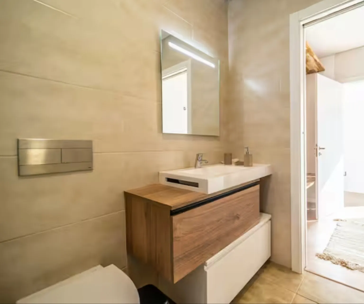 Baño en tonos arena con mueble de madera, espejo retroiluminado y puerta abierta