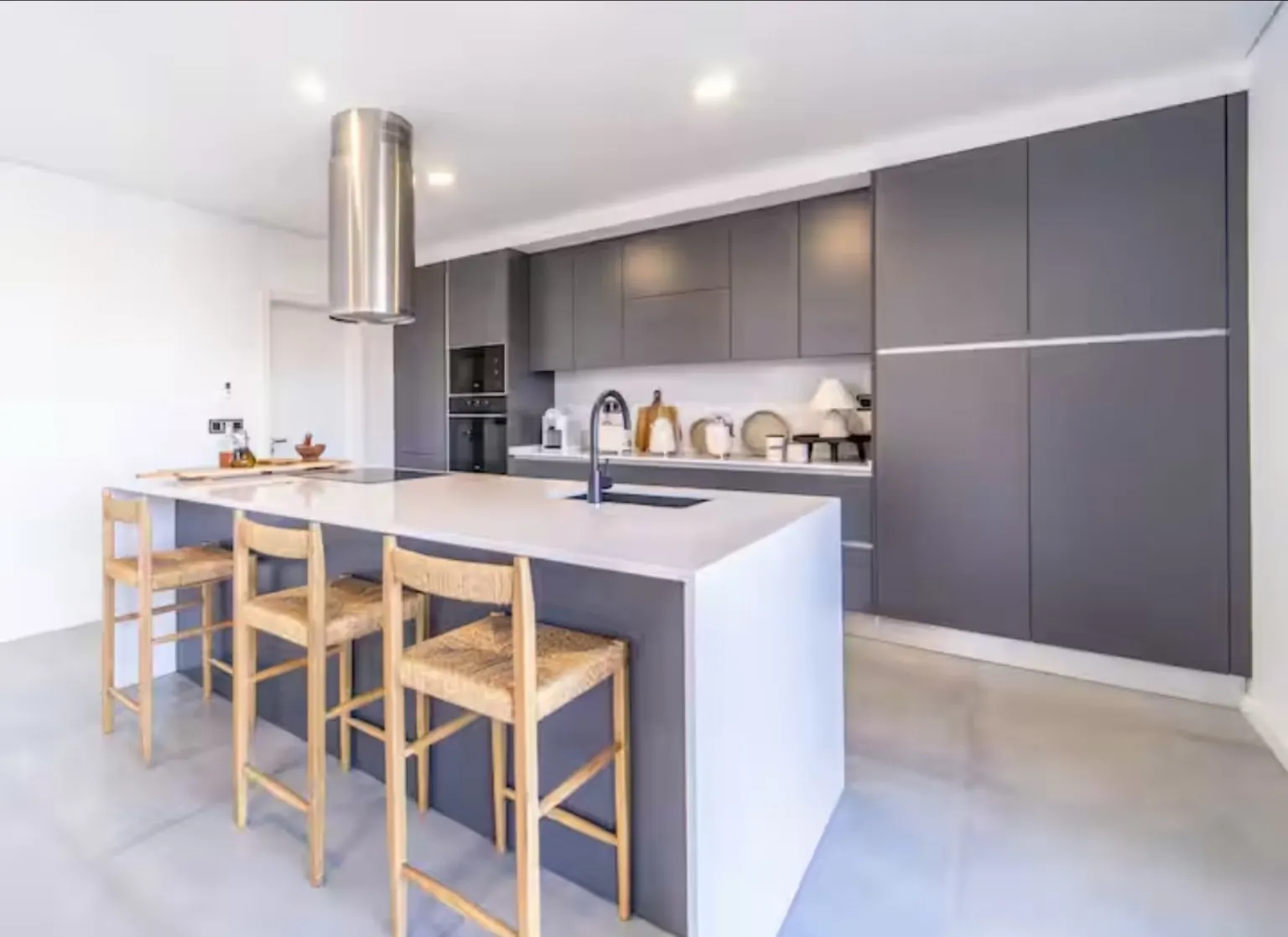 La misma cocina gris tras la reforma, con taburetes de madera y enea y accesorios naturales en la encimera