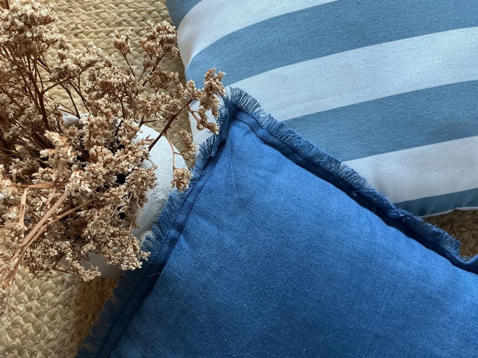Detalle de cojines de lino azul y rayas junto a flores secas en una cesta de fibra