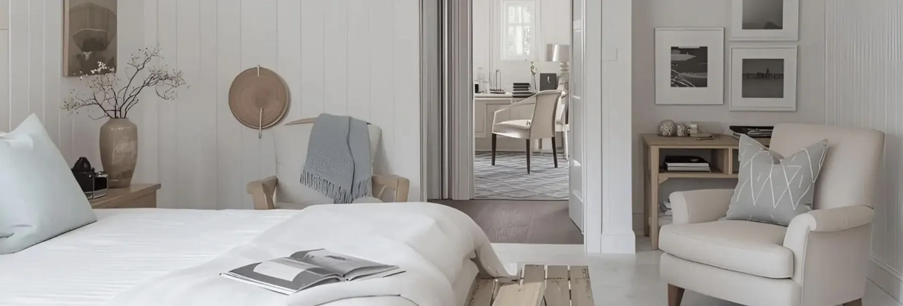 Dormitorio en tonos claros con cama de lino, butaca tapizada y puerta abierta a un pasillo luminoso