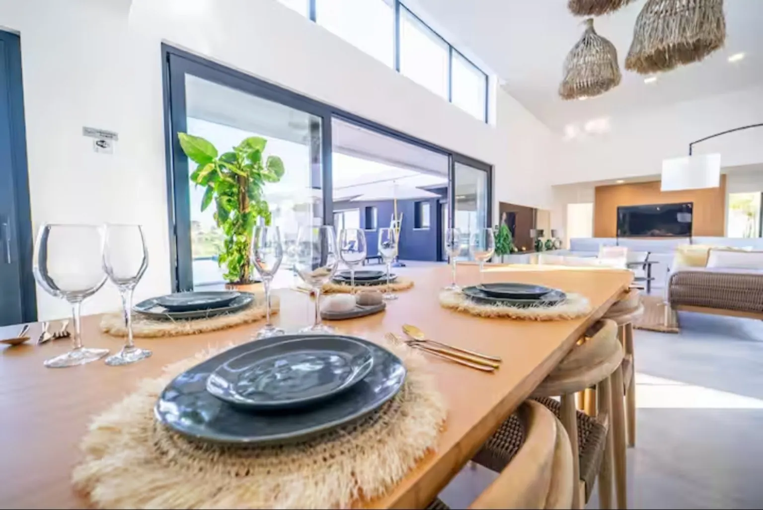 Mesa de comedor de madera con bajoplatos de fibra, platos azul oscuro y lámparas de mimbre junto a un ventanal