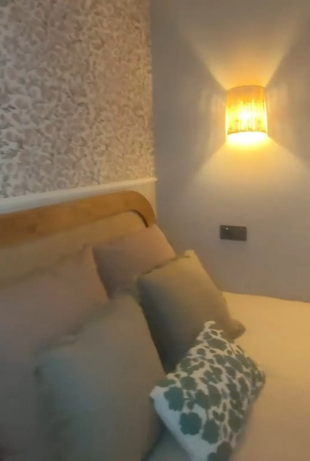 Detalle de cama con cojines estampados bajo un aplique de luz cálida y papel floral