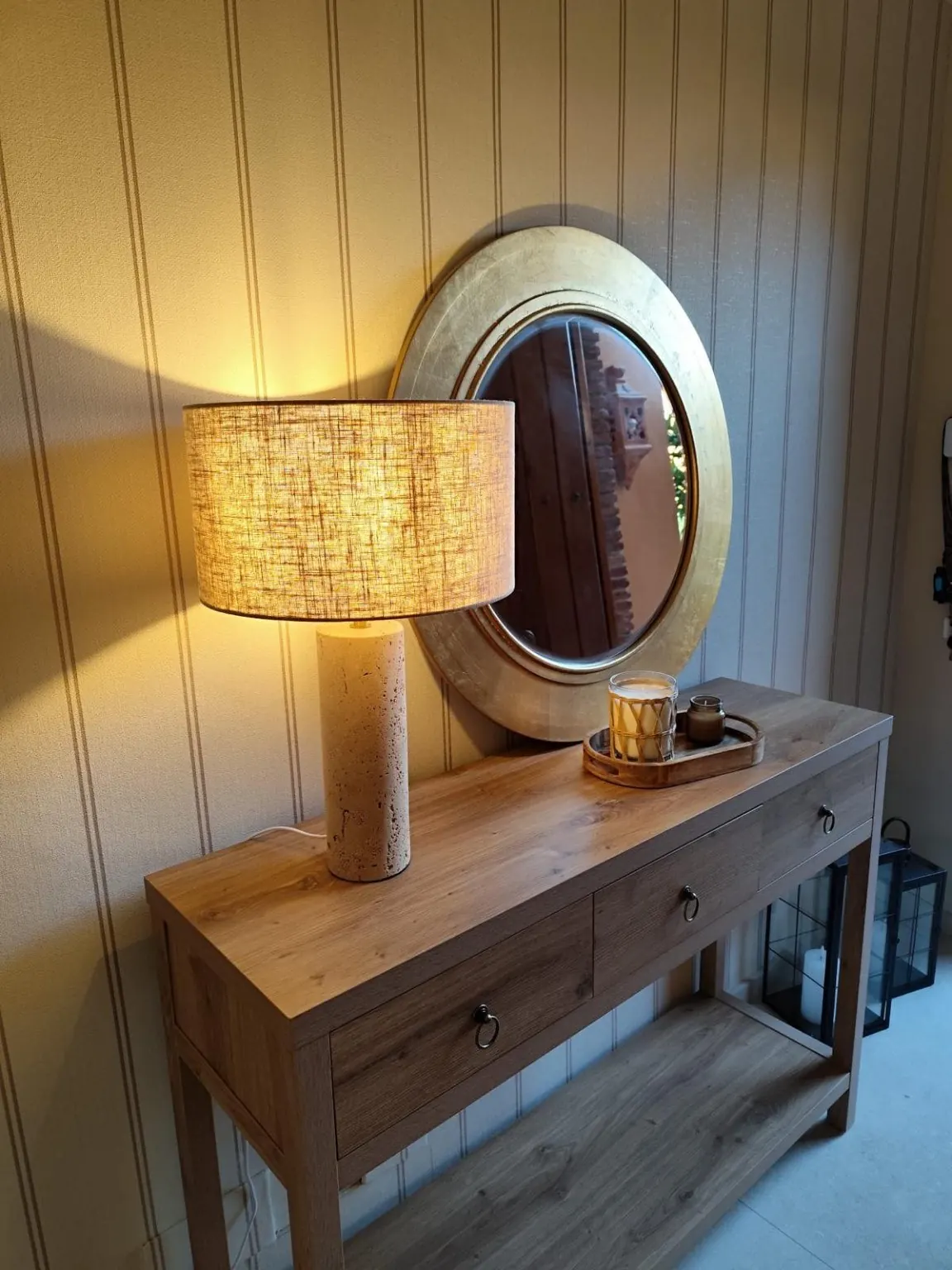 Recibidor con consola de madera, lámpara de lino encendida y espejo redondo dorado