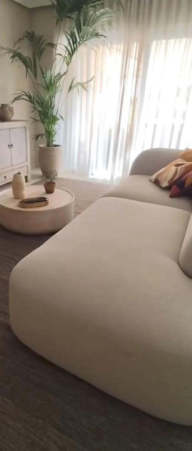 Sofá modular beige junto a una ventana con cortinas claras y una palmera en maceta